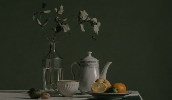 A dark still life of a teapot, jug and fruit on a table, lit from one side.