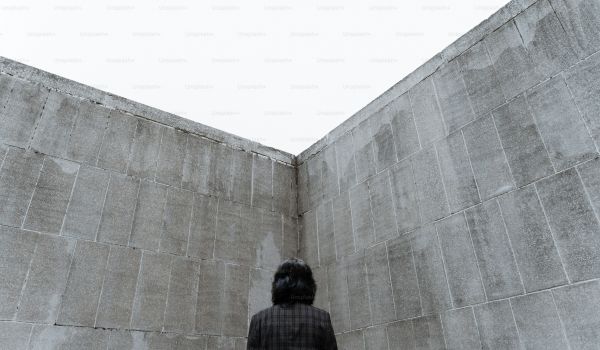 A person standing in the corner of two high concrete walls, seen from behind, looking up.