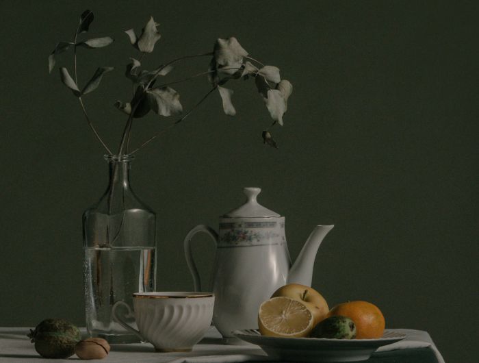 A dark still life of a teapot, jug and fruit on a table, lit from one side.