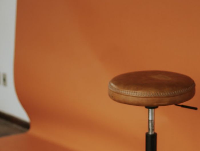 A leather stool standing empty in front of an orange studio backdrop, with a lighting stand to one side