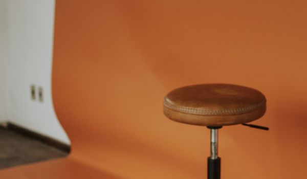A leather stool standing empty in front of an orange studio backdrop, with a lighting stand to one side