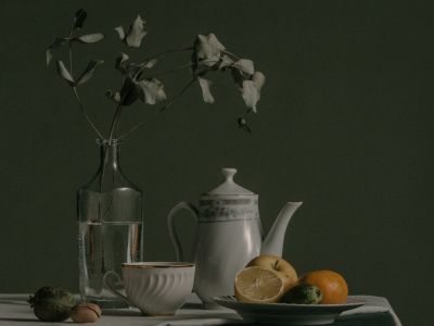A dark still life of a teapot, jug and fruit on a table, lit from one side.