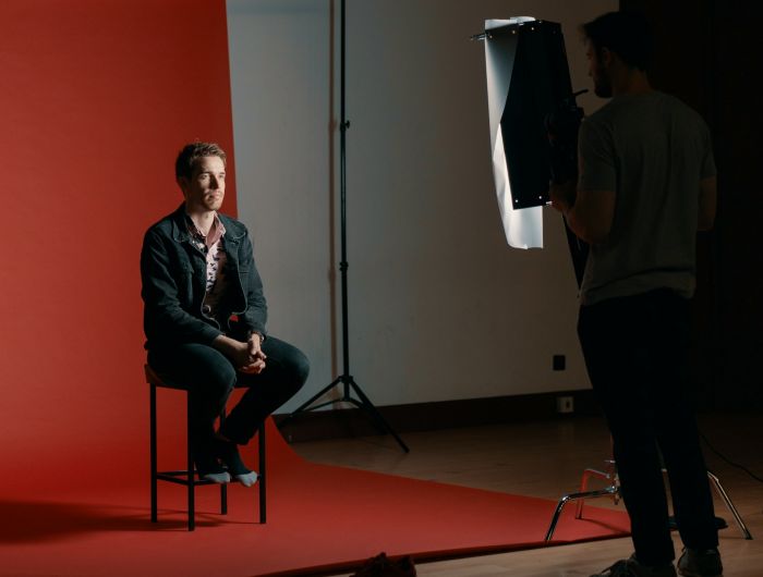 A man sitting on a stool against a red backdrop while a crew member adjusts a light.